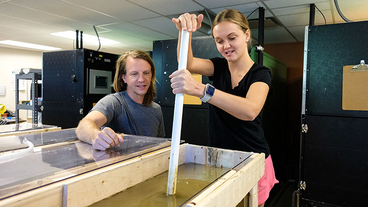 From left, Evan Guiney, associate professor of biology, guides Carmen Hay, a senior biology major, in removing a sediment core sample from one of four Wolf Creek-sourced mesocosms in a lab at SRU. The pair will later identify and sequence microbe DNA from the samples and analyze its potential for pollution remediation.