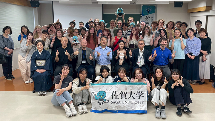 Group of students and faculty at Saga University posing with flag and giving thumbs up.