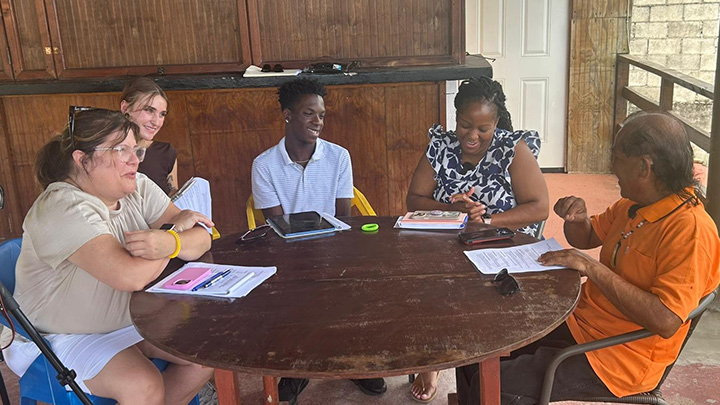 Professors and student sitting at a table interviewing a member of the Kalinago community.