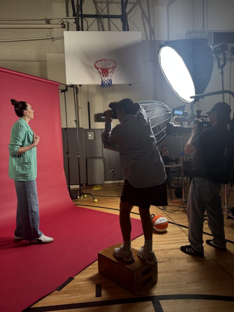 Photographer shooting a basketball player on a court with light fixtures and backdrop.