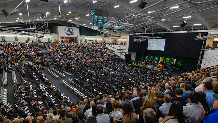 Wide angle view of Morrow Field House commencement stage with graduates in their seats and spectators in the bleachers.