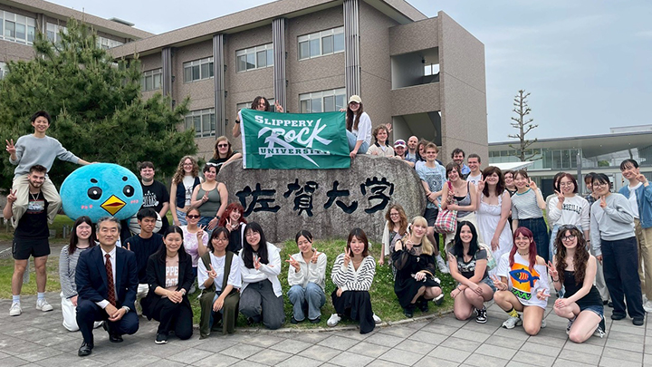 Group of 30 students and professors standing outside a building with SRU flag.