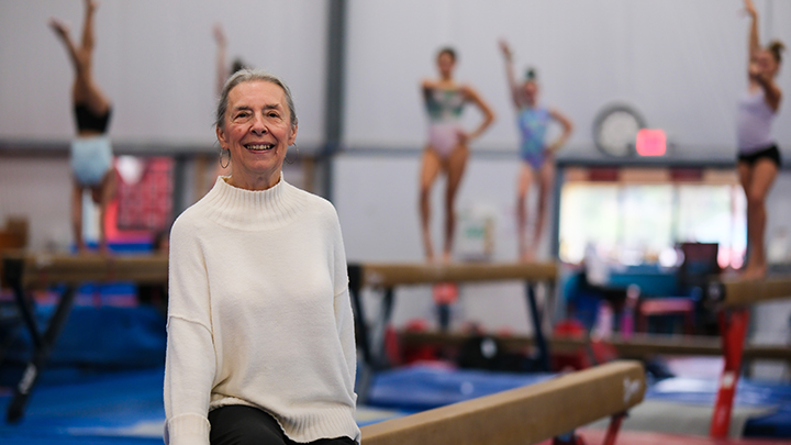 Woman standing in front of a balance beam with gymnasts in background.