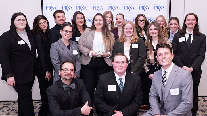 Group photo of students, faculty and alumni holding a trophy in front of a PRSA backdrop.