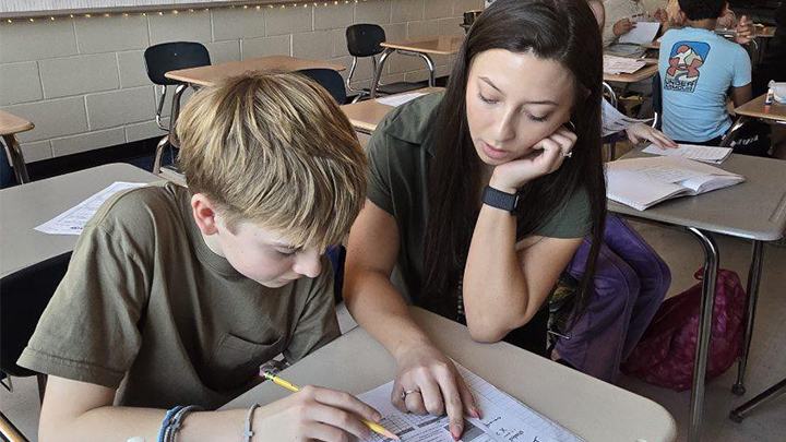 Teacher instructing a student who is writing while sitting at a desk.