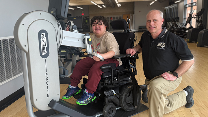 Person using a wheelchair operating an arm ergometer with another person kneeling next to him.