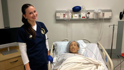 Nursing standing next to hospital bed with simulation patient laying in it.