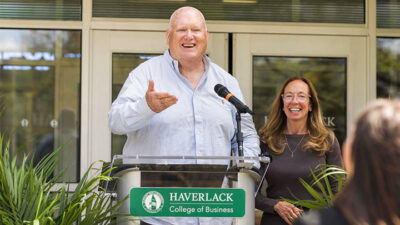 Man speaking at a podium with woman smiling in background.