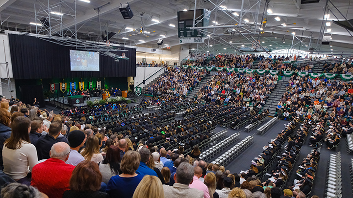 Wide view of commencement stage inside the gym with bleachers filled with spectators and floor seats filled with graduates.