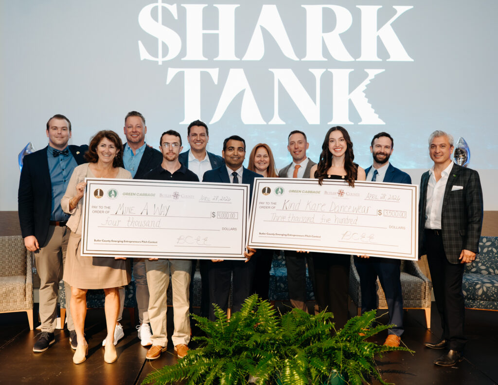 Two people holding giant checks while surrounding by nine other people with a sign that says Shark Tank in the background.
