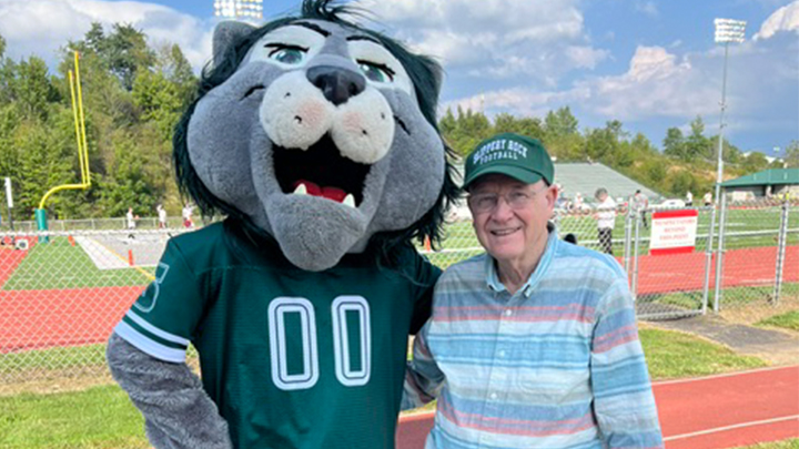 Rocky lion mascot standing next to man in SRU hat at a football field.