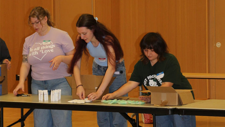 Three students standing at a table assembling packages of hygiene products.