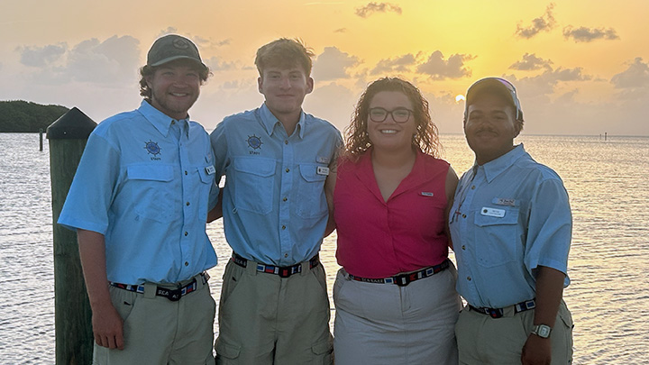 Four people standing side by side in front of an ocean sunset.