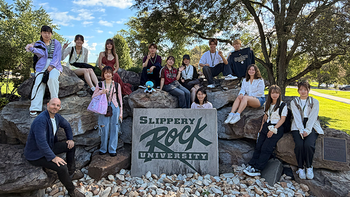 More than a dozen students and faculty sitting around a rock formation near a sign that says Slippery Rock.