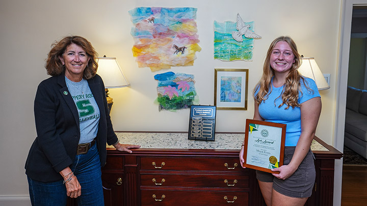 From left, SRU President Karen Riley and senior art major Megan Evans stand in the foyer of the President’s Residence next to Evans’ artwork. Evans was selected as the winner of the President’s Student Art in Residence Award for Fall 2026.