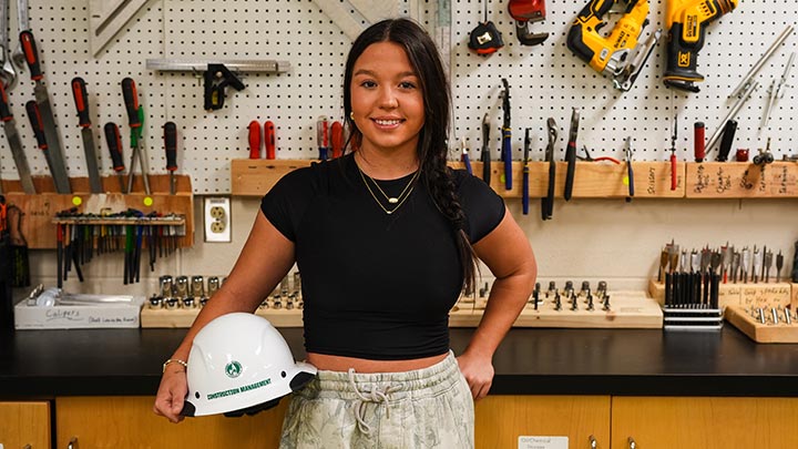 Woman holding a hardhat standing in construction lab with tools on wall.