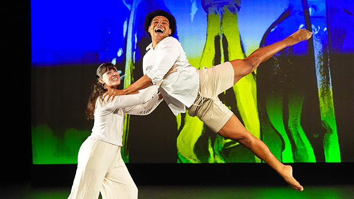 One dancer holding up another dancer on stage with art in the background.